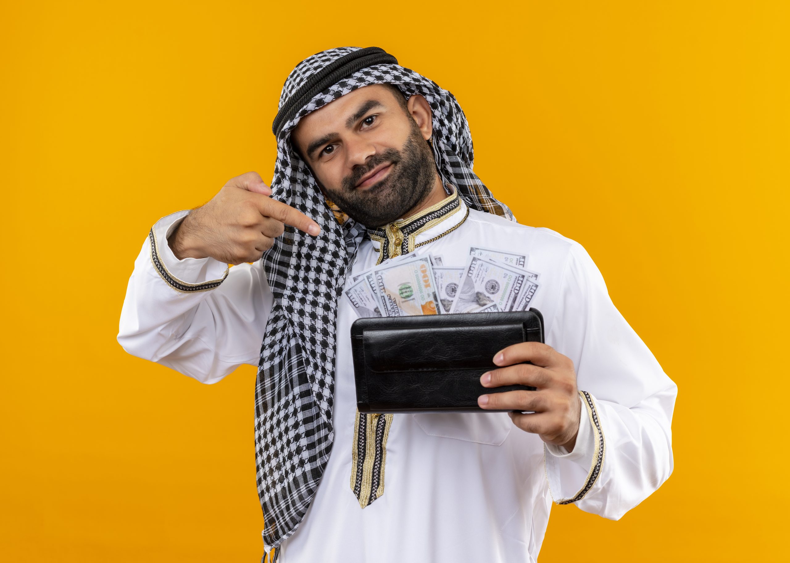 arabic businessman in traditional wear holding wallet with cash looking at camera smiling confident pointing with finger to cash standing over orange background